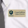 Close-up of a professional name tag worn on a white dress shirt with a gray tie. The name tag is gold with black text, displaying 'Liam Jeffries, Adjunct Faculty' alongside a College logo featuring a lion emblem.