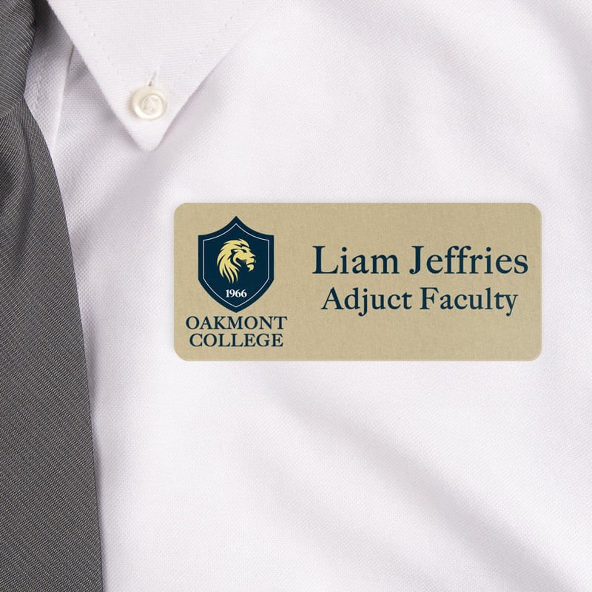 Close-up of a professional name tag worn on a white dress shirt with a gray tie. The name tag is gold with black text, displaying 'Liam Jeffries, Adjunct Faculty' alongside a College logo featuring a lion emblem.