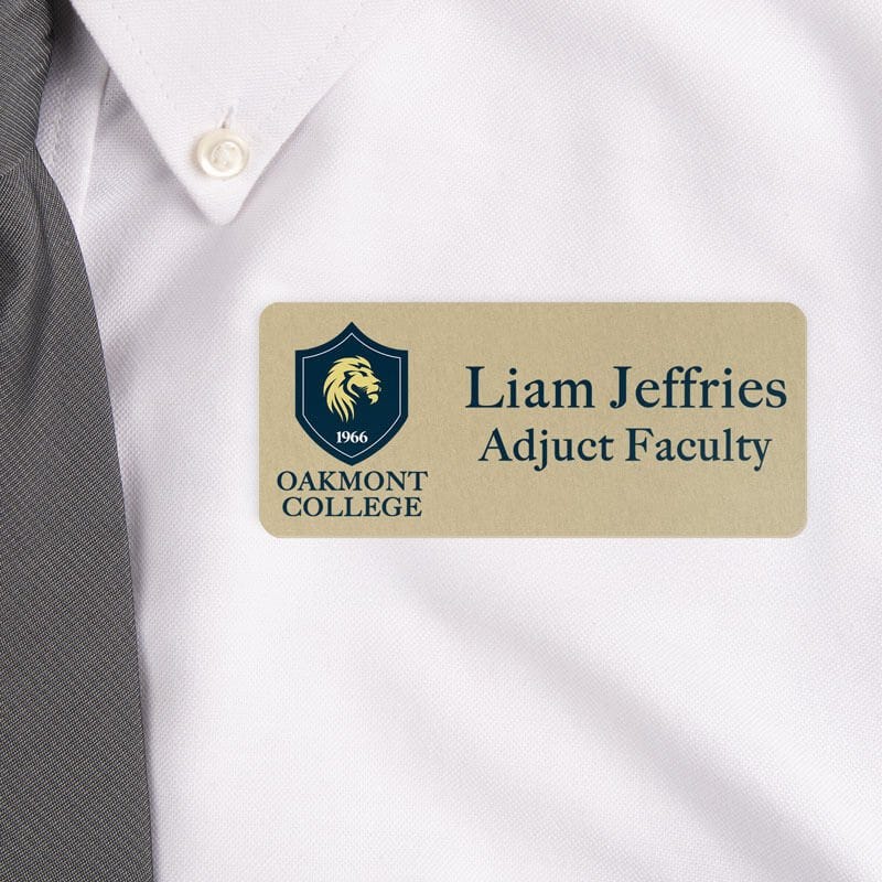 Close-up of a professional name tag worn on a white dress shirt with a gray tie. The name tag is gold with black text, displaying 'Liam Jeffries, Adjunct Faculty' alongside a College logo featuring a lion emblem.